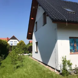 Nowoczesny dom jednorodzinny z białą elewacją i ciemną dachówką, widok z ogrodu w słoneczny dzień, okno zabezpieczone folią ochronną.