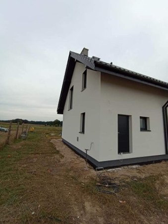 Świeżo otynkowany, biały dom jednorodzinny z czarnymi detalami (okna, drzwi, dach) na tle pochmurnego nieba i niezagospodarowanego terenu.