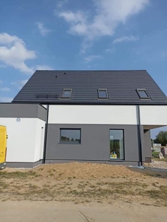 Nowoczesny dom jednorodzinny z szarą elewacją i ciemnoszarą dachówką, widoczne trzy okna dachowe, teren wokół domu częściowo zagospodarowany, z widocznym piaskiem.