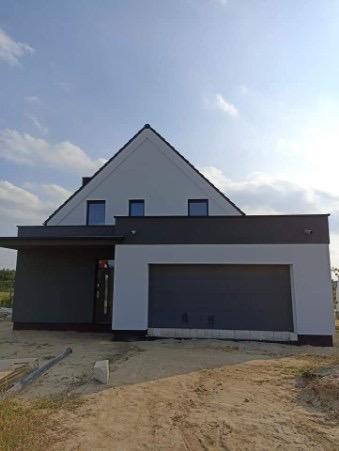 Nowoczesny dom jednorodzinny z dwukolorową elewacją: biała góra i grafitowy dół, widok na front z bramą garażową i trzema oknami na poddaszu, w tle błękitne niebo.