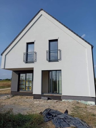 Nowoczesny, dwukondygnacyjny dom z białymi ścianami i szarymi ramami okiennymi, widoczne małe balkony z metalowymi barierkami, teren wokół domu jeszcze niezagospodarowany, z resztkami materiałów...