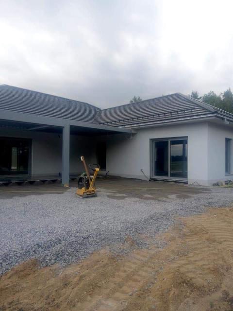 Niedokończona elewacja domu jednorodzinnego z szarym dachem, widoczna ubijarka wibracyjna na podłożu z kamieni, przygotowanie terenu przed budynkiem.