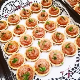 Taca z tartaletkami wypełnionymi kremowym musem, udekorowanymi świeżymi ziołami, ułożonymi na ażurowej serwetce.