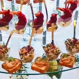 Elegancki catering: Różnorodne przystawki w szklanych naczyniach, w tym krewetki w tempurze na szpadce, sałatki z kawiorem i roladki z łososia, ułożone na szklanym, wielopoziomowym stojaku.