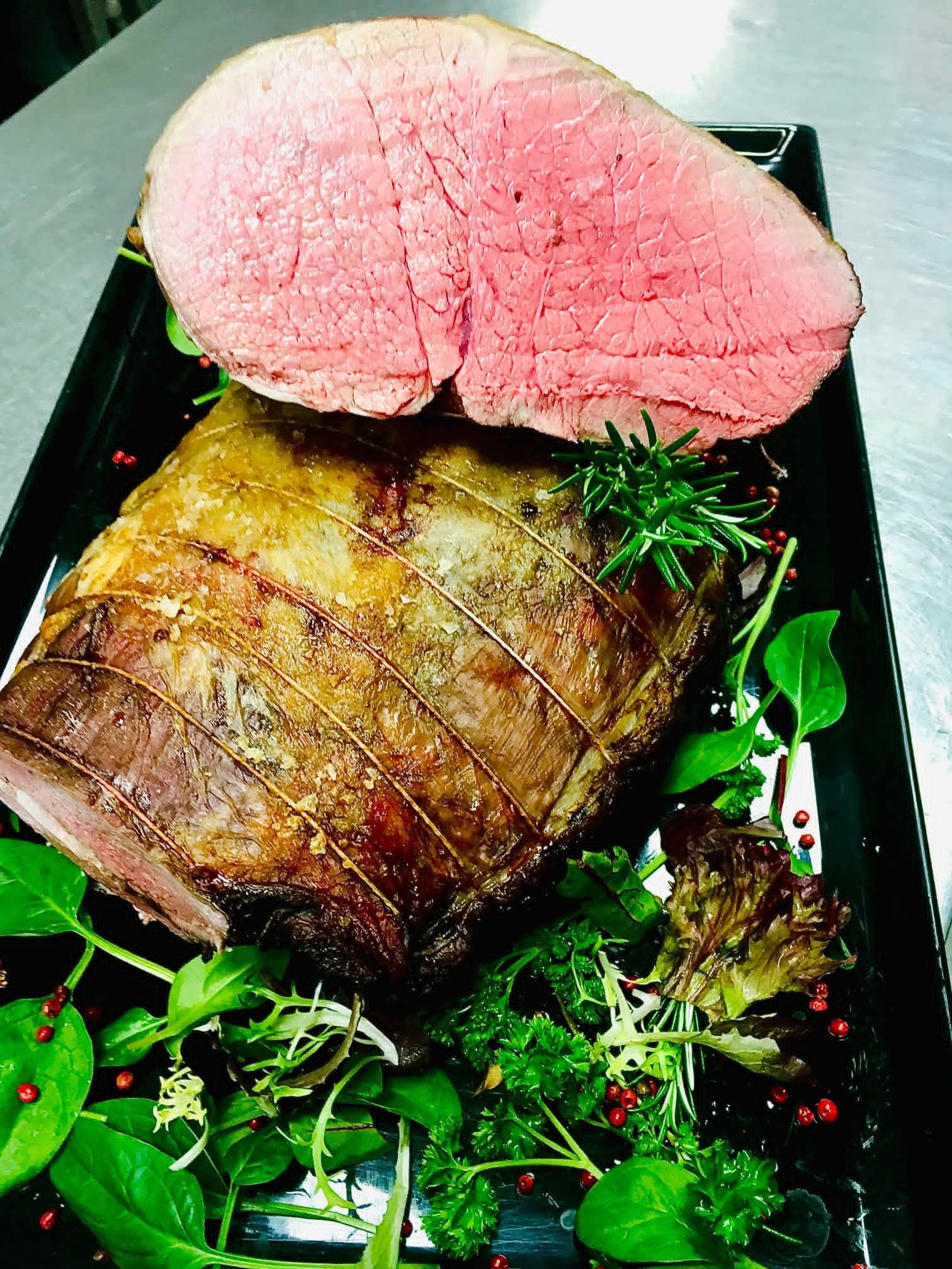 Soczysta pieczeń wołowa na czarnym talerzu, udekorowana świeżymi ziołami i zielonymi liśćmi. Mięso jest różowe w środku, a skórka ma złocisty kolor.