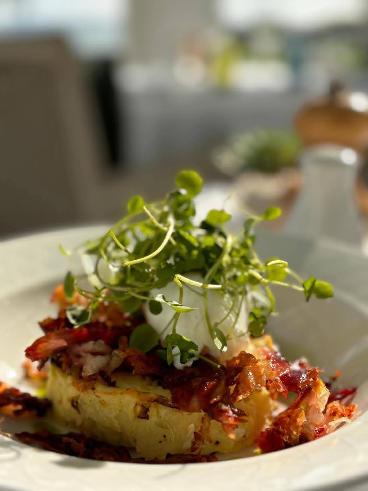 Apetyczna kompozycja: ziemniaczany placek z chrupiącym boczkiem, delikatnym jajkiem w koszulce i świeżymi mikrolistkami na białym talerzu w jasnym otoczeniu.