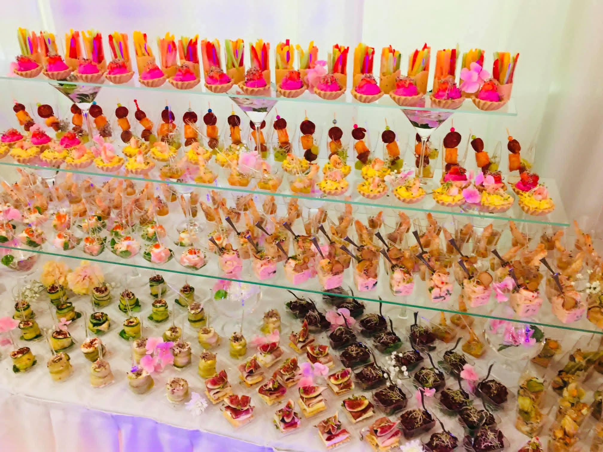 Bogato zastawiony stół z przekąskami na kilku poziomach, dekorowany kwiatami. Różnorodność kanapek, tartaletek i szaszłyków tworzy apetyczną kompozycję.