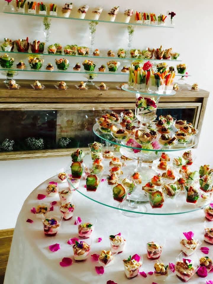 Elegancki stół cateringowy z różnorodnymi przekąskami w szklanych naczyniach, dekorowany płatkami róż, z półkami pełnymi finger food w tle.