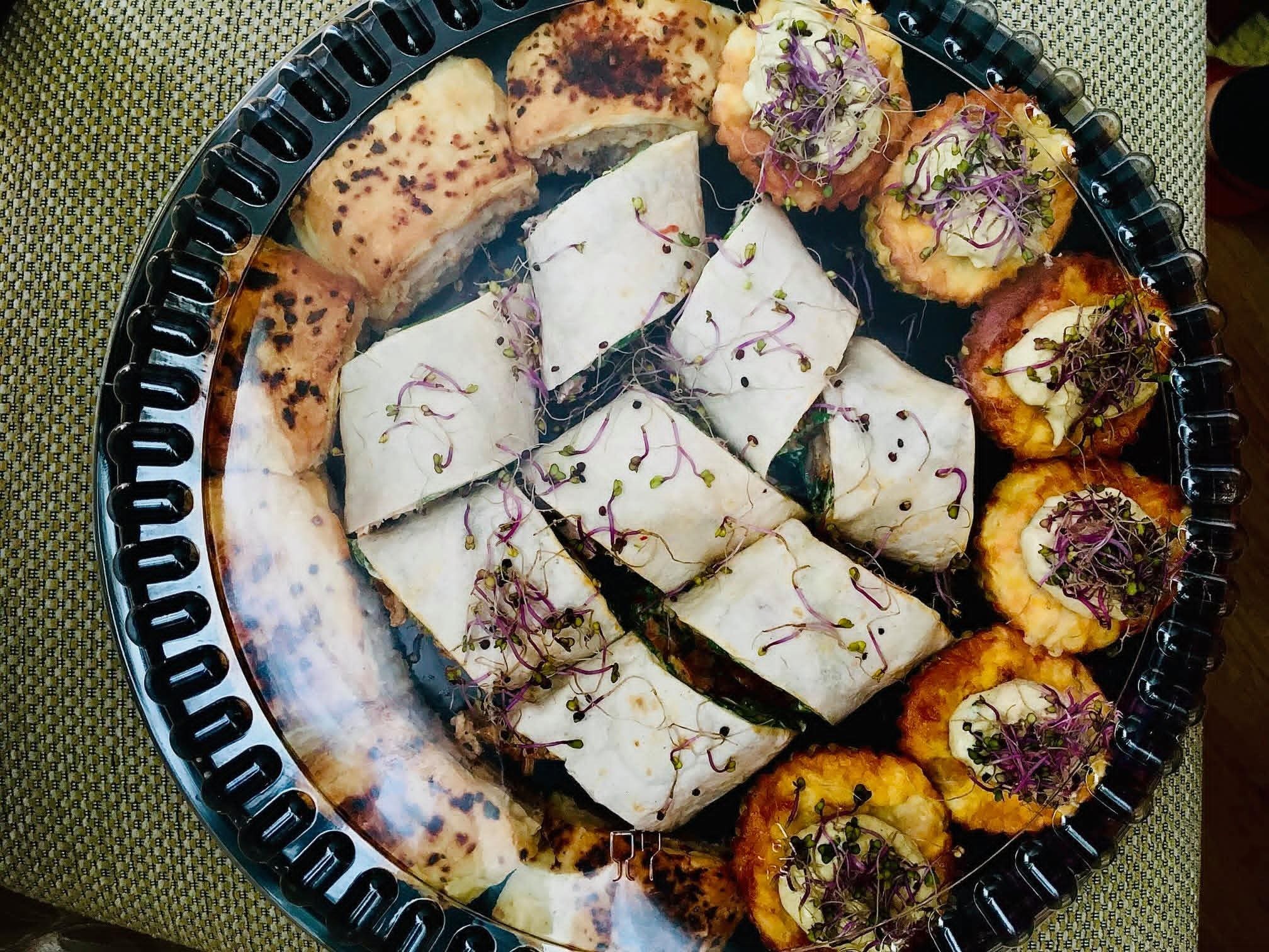 Elegancka taca z różnorodnymi przekąskami: roladki z kiełkami, tartaletki z kremem i ziołami, oraz pieczywo. Całość w czarnej, przezroczystej tacy.