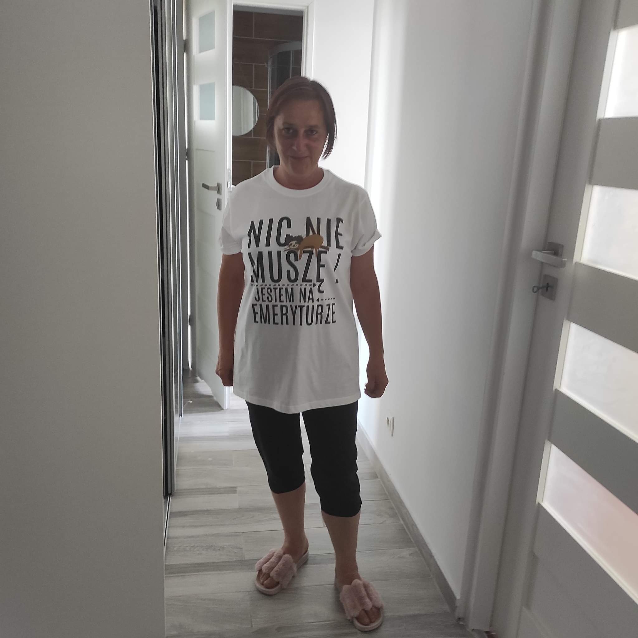 Kobieta w koszulce z napisem 'Nic nie muszę! Jestem na emeryturze' i różowych kapciach stoi w korytarzu z widocznymi drzwiami do łazienki z okrągłym lustrem.