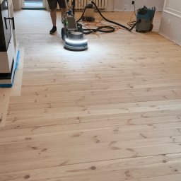 PAN CYKLINIARZ - Rzetelny Wykonawca - Maszyna do szlifowania podłogi w trakcie pracy, widoczny operator, odkurzacz przemysłowy i fragment świeżo wyszlifowanej drewnianej podłogi z jasnego drewna.