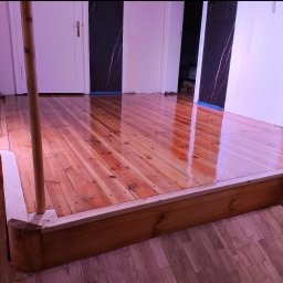 PAN CYKLINIARZ - Rzetelny Wykonawca - Lakierowana podłoga drewniana, częściowo podwyższona, z widocznymi śladami świeżo nałożonego lakieru i zabezpieczoną taśmą malarską przy ścianach i drzwiach, podczas aplikacji lakieru wałkiem.