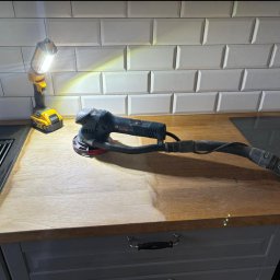 PAN CYKLINIARZ - Rzetelny Wykonawca - Szlifowanie drewnianego blatu kuchennego szlifierką mimośrodową Bosch, z widocznym fragmentem blatu przed i po obróbce, oświetlone lampą roboczą DeWalt.