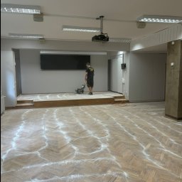 PAN CYKLINIARZ - Rzetelny Wykonawca - Duża sala z parkietem w jodełkę częściowo pokrytym pyłem po szlifowaniu, pracownik w czarnej koszulce obsługuje cykliniarkę na podwyższeniu, widoczny projektor sufitowy i oświetlenie jarzeniowe.
