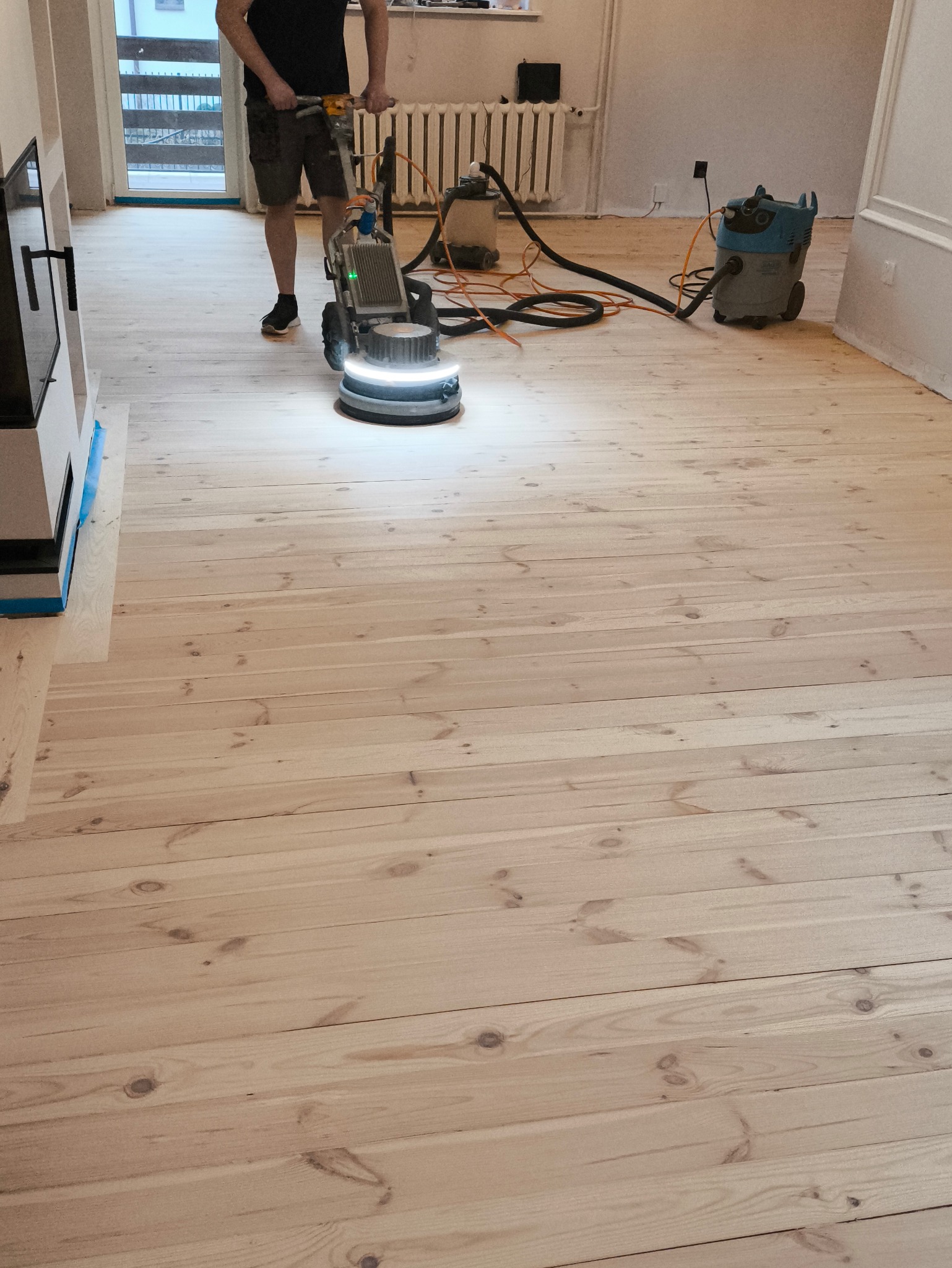 Maszyna do szlifowania podłogi w trakcie pracy, widoczny operator, odkurzacz przemysłowy i fragment świeżo wyszlifowanej drewnianej podłogi z jasnego drewna.