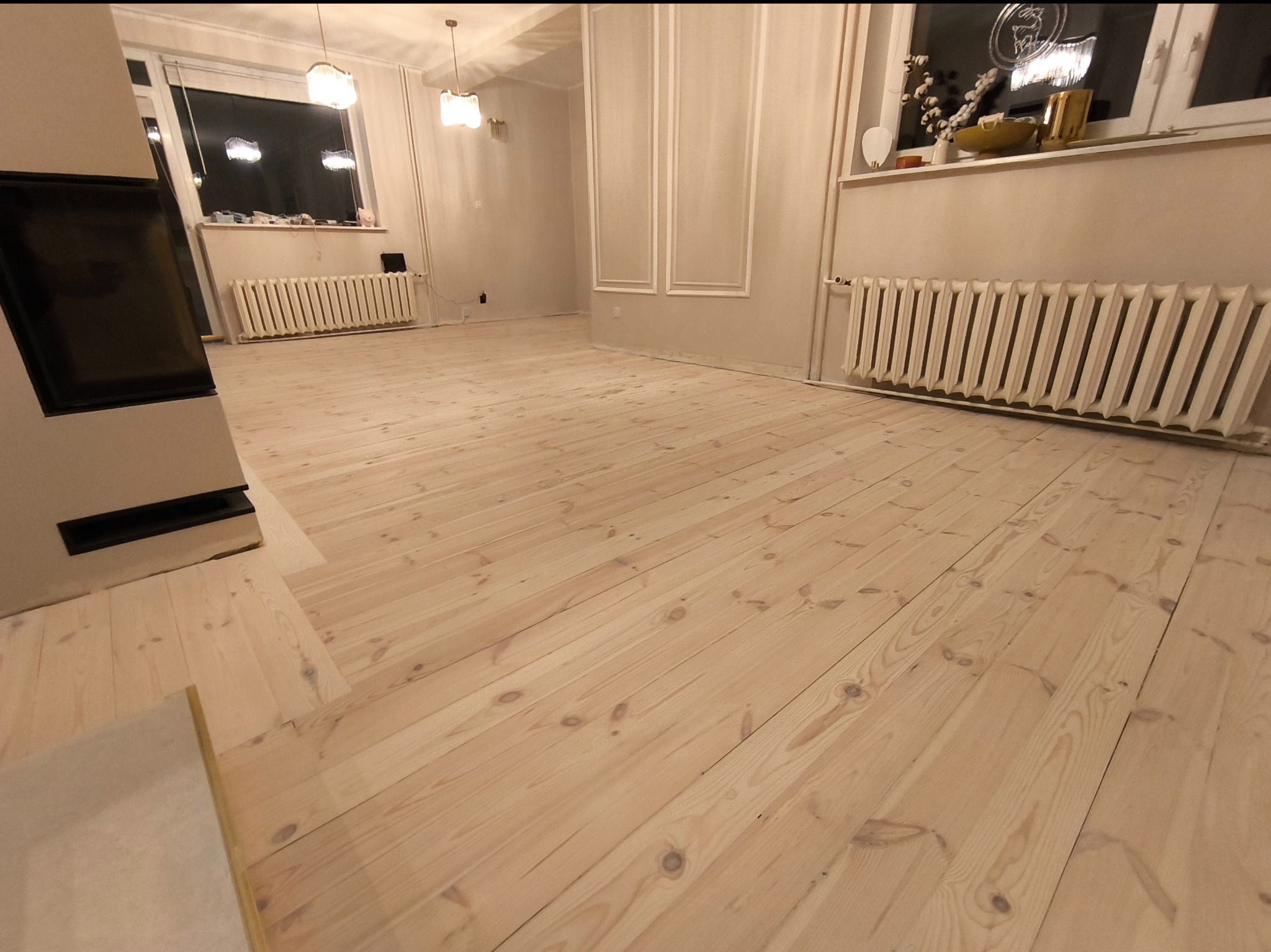 Jasne, odnowione drewniane deski podłogowe ułożone w jodełkę w przestronnym, jasnym pomieszczeniu z grzejnikami pod oknami i dekoracyjnymi listwami na ścianach.