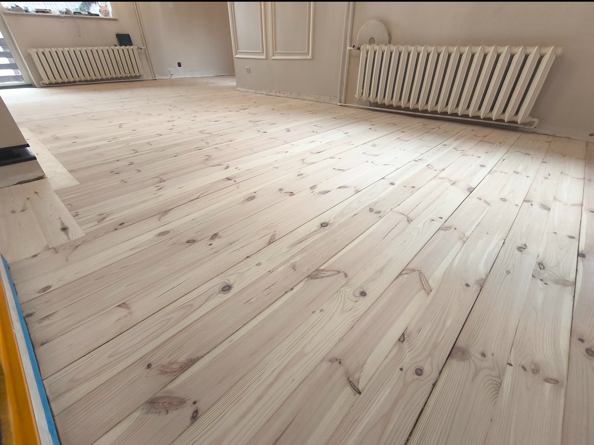 Jasne, świeżo wycyklinowane deski podłogowe z widocznym rysunkiem drewna i białym olejem, w tle grzejnik żeberkowy i fragment ściany z dekoracyjnymi listwami.