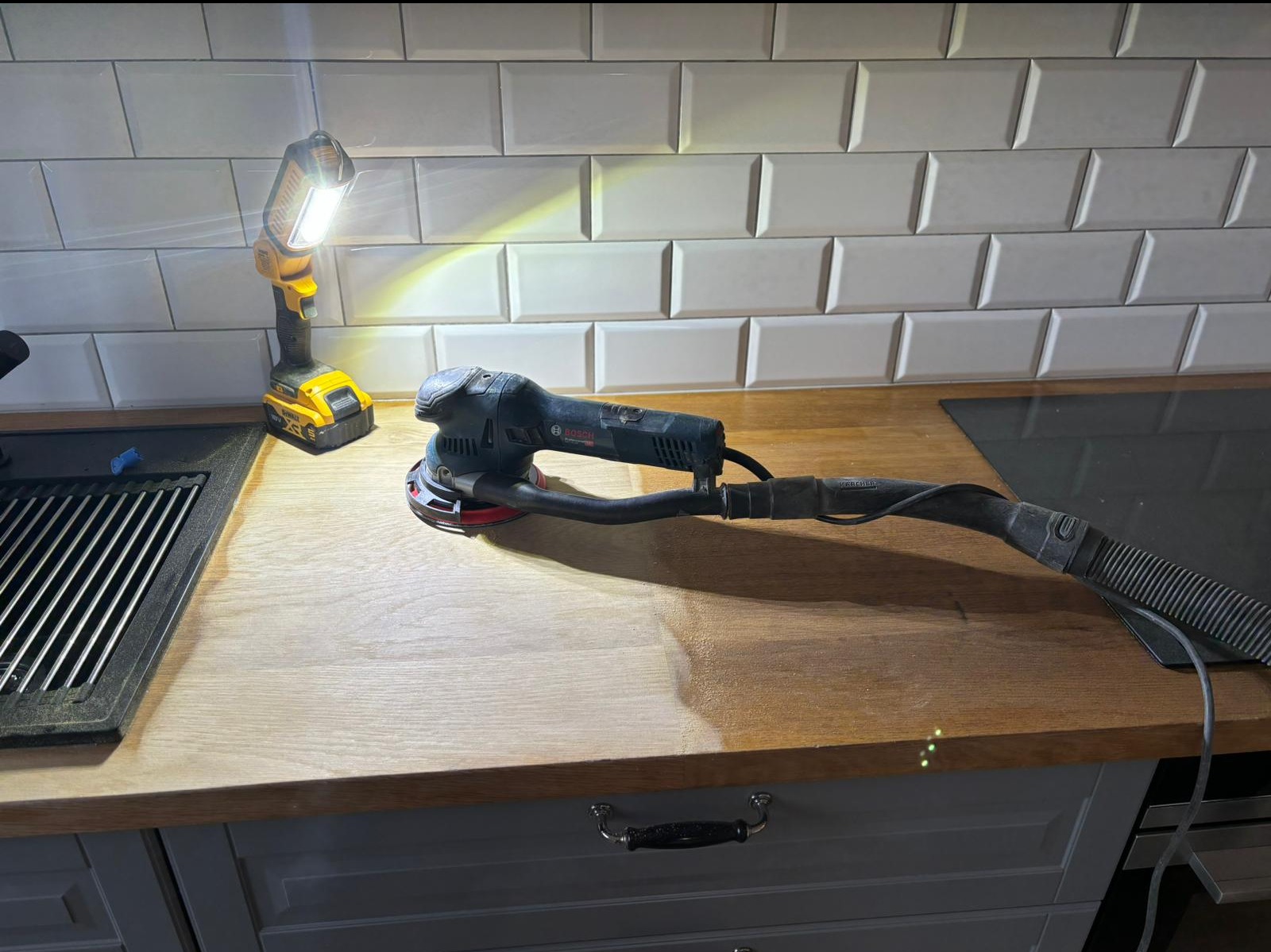 Szlifowanie drewnianego blatu kuchennego szlifierką mimośrodową Bosch, z widocznym fragmentem blatu przed i po obróbce, oświetlone lampą roboczą DeWalt.