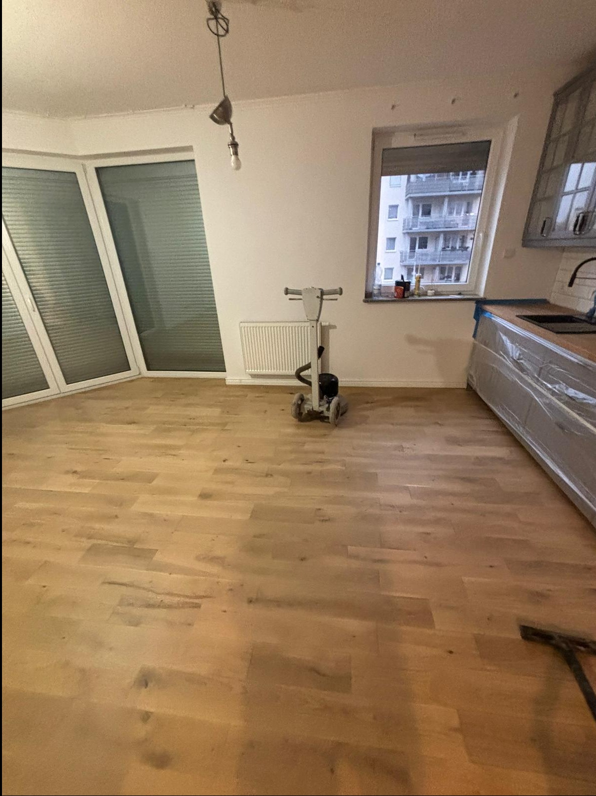Maszyna do renowacji parkietu stoi na wycyklinowanej drewnianej podłodze w jasnym pokoju z dużymi oknami balkonowymi i osłoniętym folią blatem kuchennym.