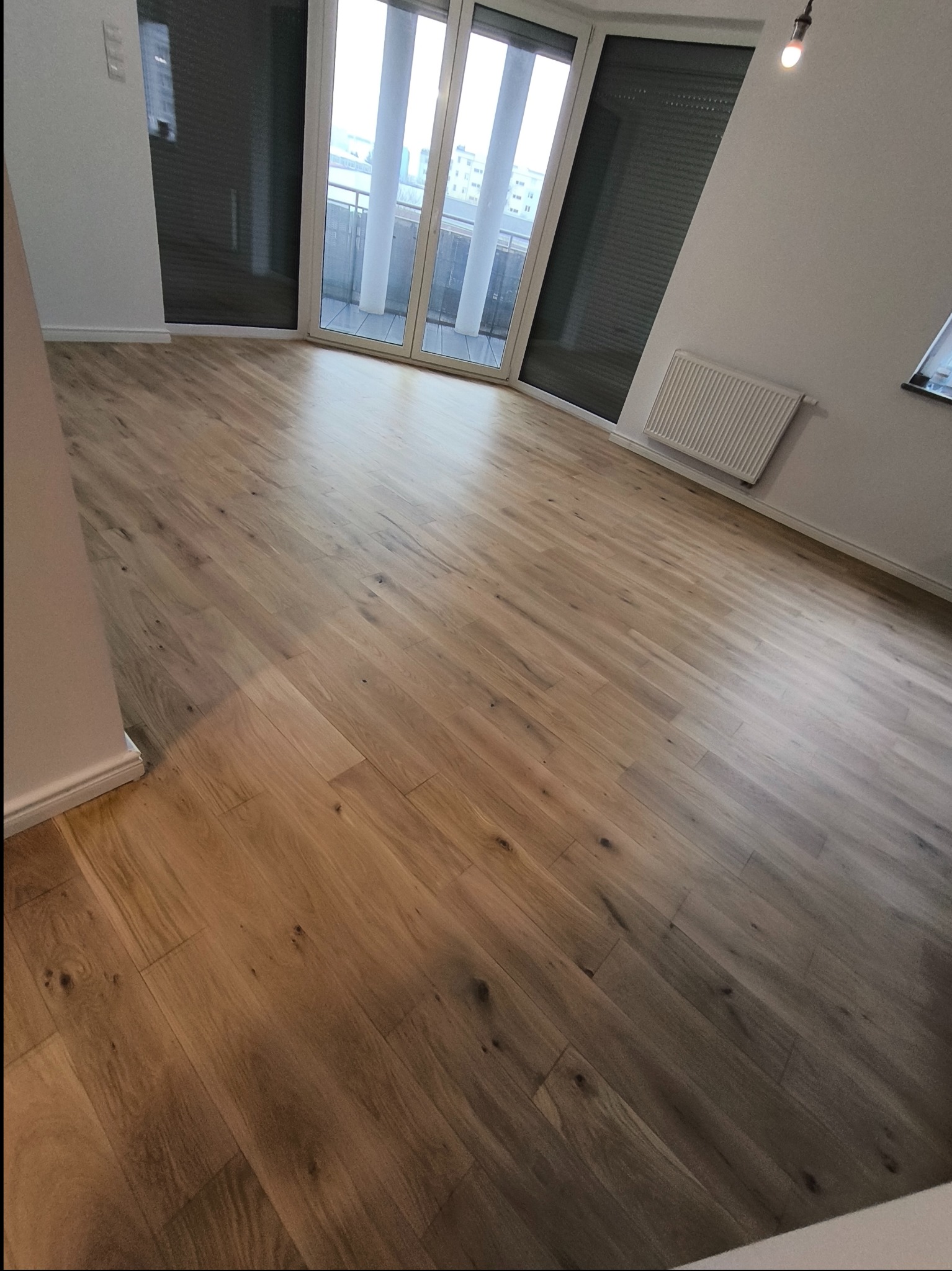 Wycyklinowana podłoga drewniana w jasnym odcieniu dębu, po renowacji, widok na balkon z dużymi oknami, minimalistyczne wnętrze z grzejnikiem na ścianie.