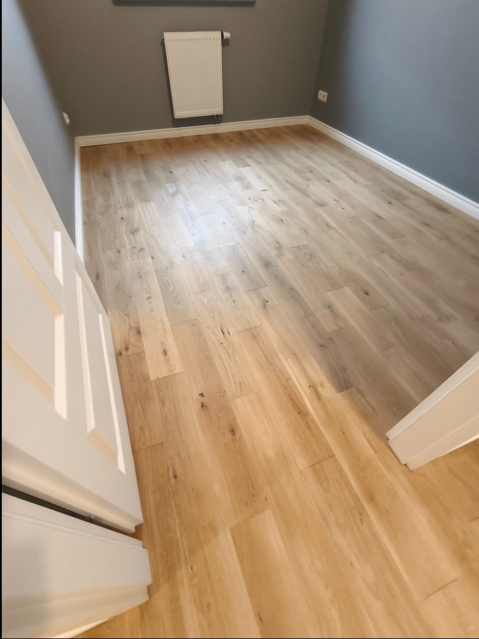 Jasna, drewniana podłoga po renowacji w pokoju z szarymi ścianami i białymi listwami przypodłogowymi, widoczny fragment białych drzwi.