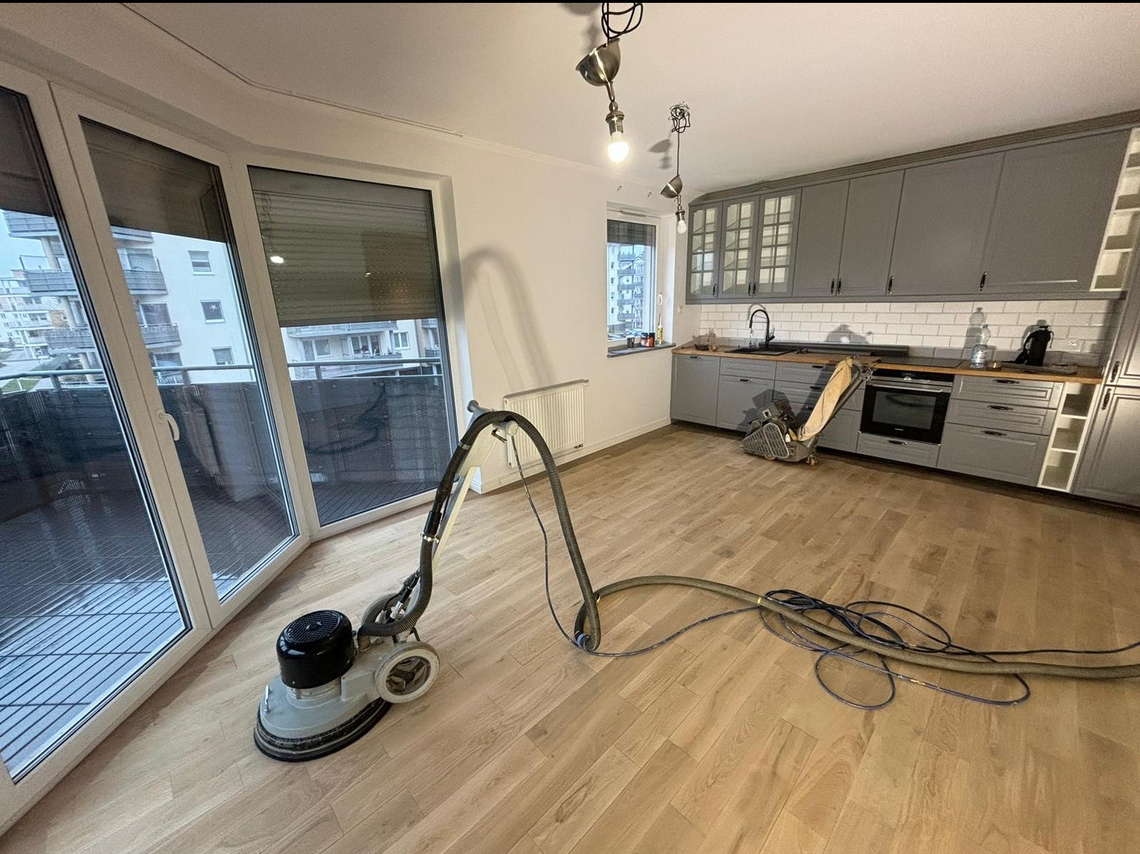 Maszyna do szlifowania parkietu w jasnym pomieszczeniu z szarymi meblami kuchennymi i dużymi oknami balkonowymi. Widoczny kabel i wąż odkurzacza. Druga maszyna stoi przy ścianie z zabudową kuchenną.