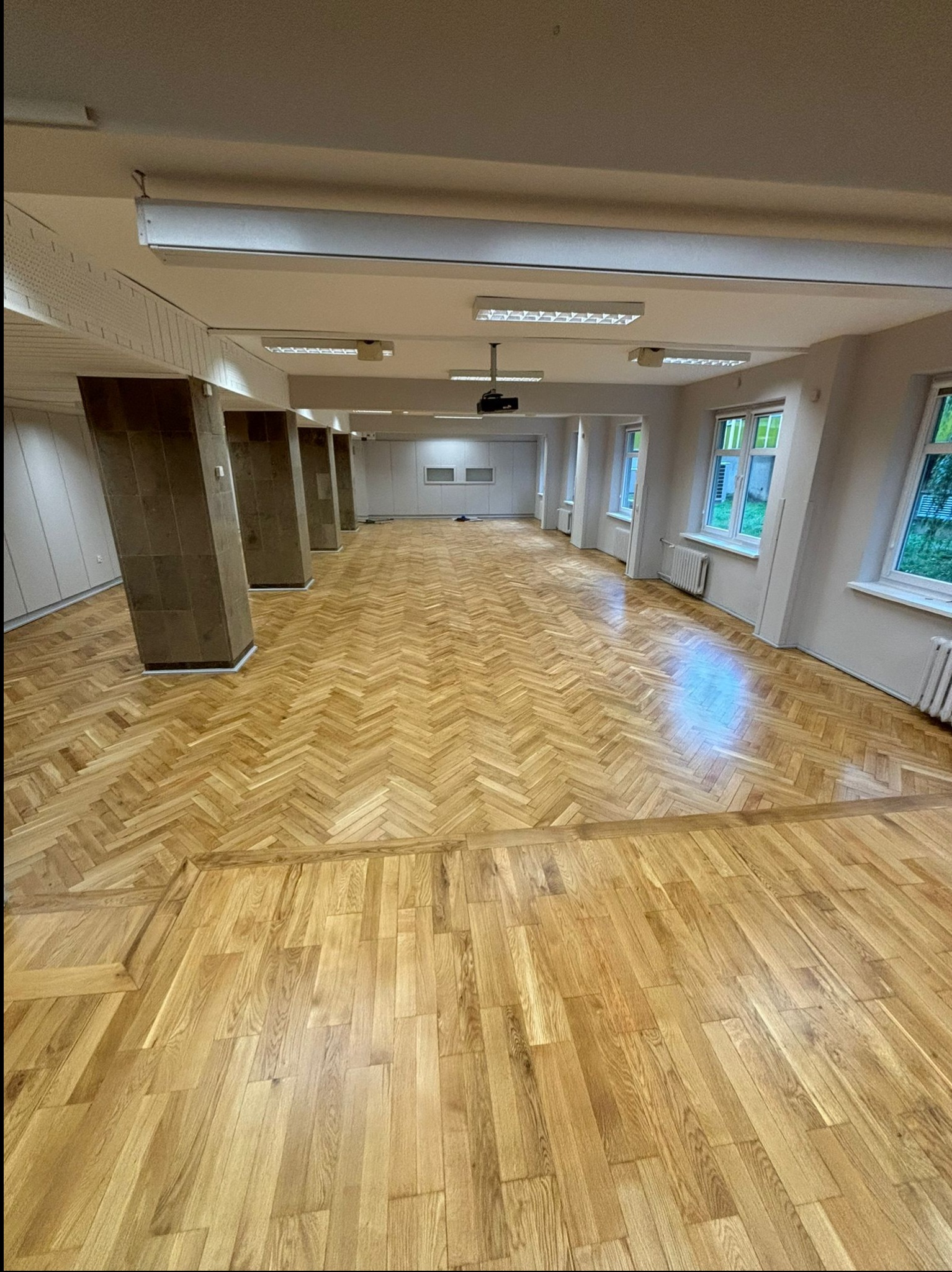 Duża sala z odnowioną, jasną parkietową podłogą w jodełkę, widoczne filary i okna z kaloryferami, rzutnik na suficie.