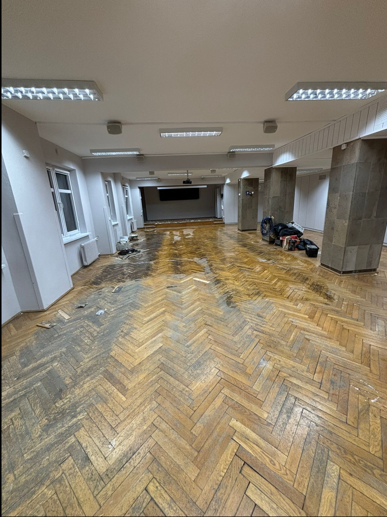 Sala z parkietem ułożonym w jodełkę w trakcie prac renowacyjnych; widoczne ślady szlifowania i zabrudzenia; w tle kolumny, okna i sprzęt do prac wykończeniowych.