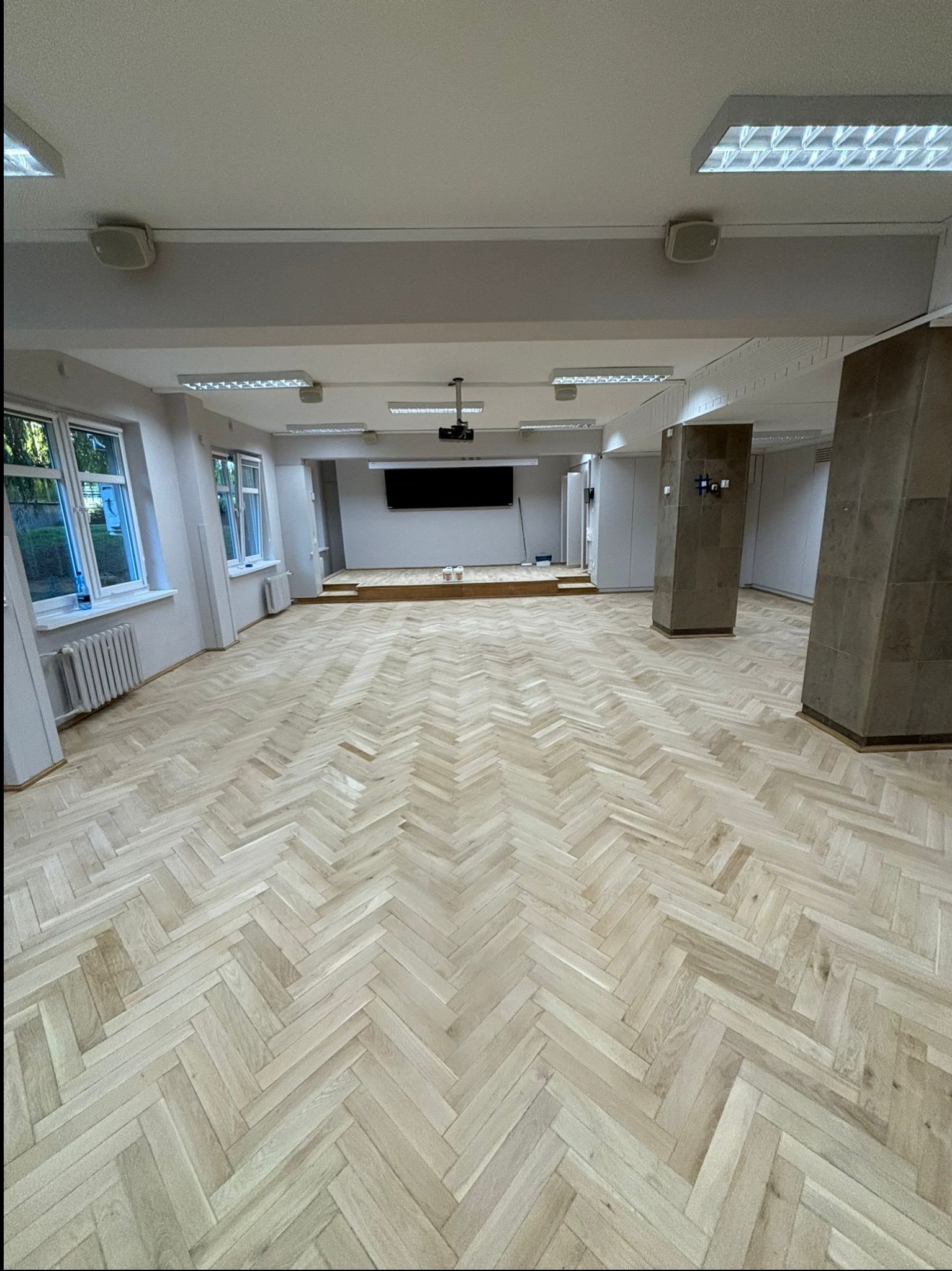 Jasna, odnowiona drewniana podłoga ułożona w jodełkę w przestronnym, jasnym pomieszczeniu z kolumnami i oknami, widoczne ślady po pracach renowacyjnych.