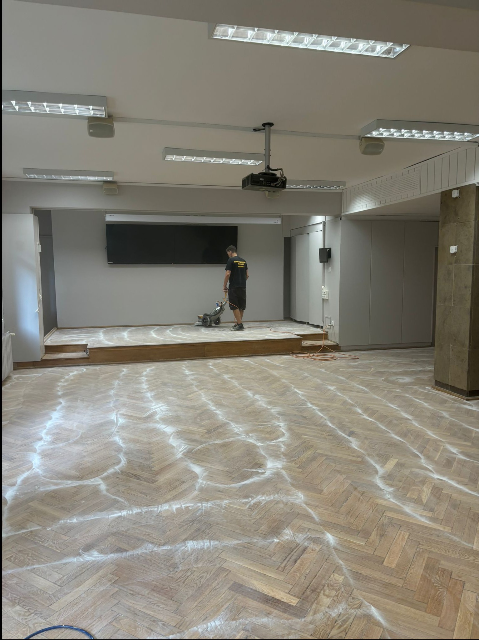 Duża sala z parkietem w jodełkę częściowo pokrytym pyłem po szlifowaniu, pracownik w czarnej koszulce obsługuje cykliniarkę na podwyższeniu, widoczny projektor sufitowy i oświetlenie jarzeniowe.