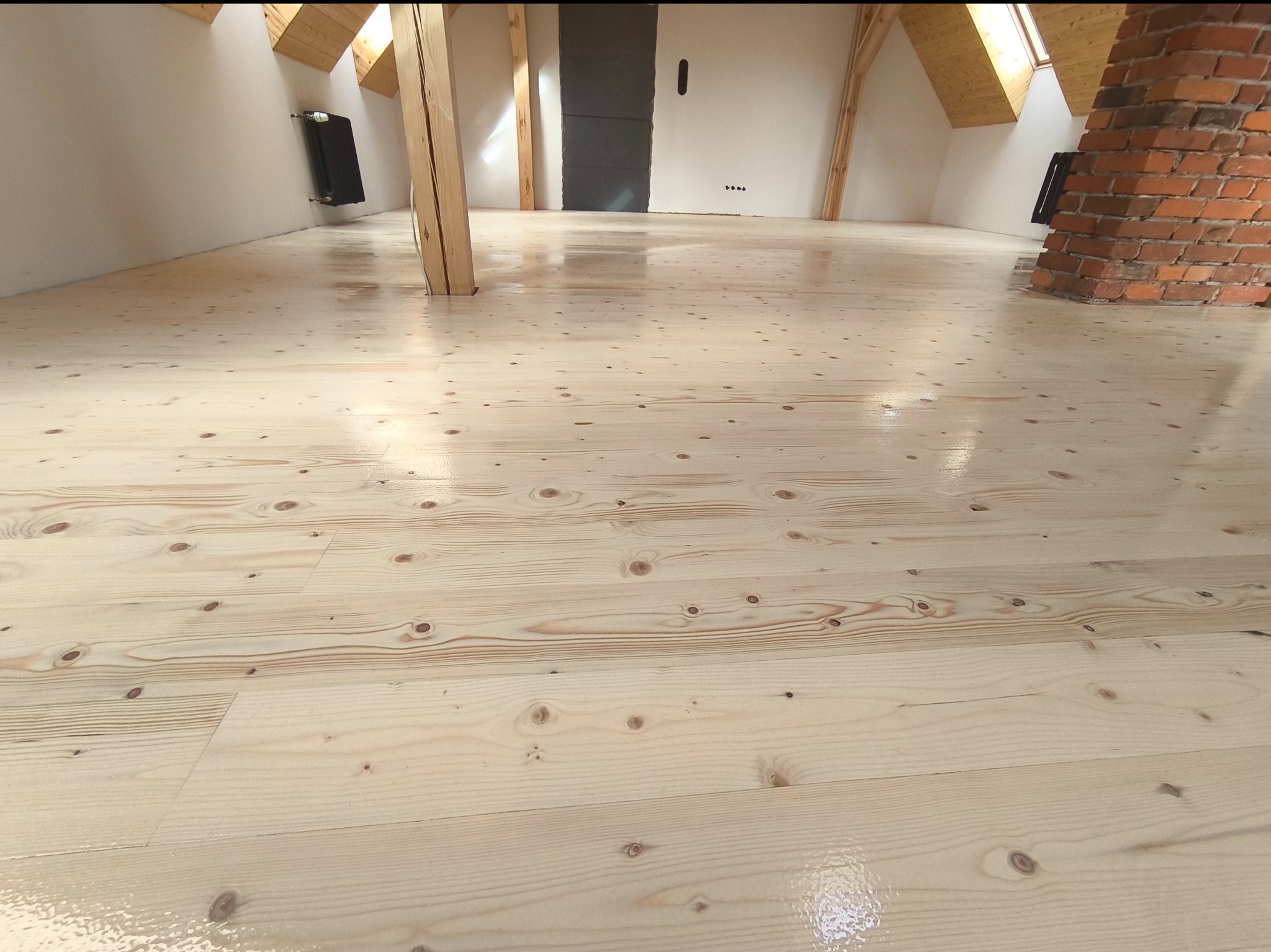 Świeżo wykończona, jasna podłoga świerkowa w pomieszczeniu na poddaszu, widoczne elementy konstrukcyjne dachu i ceglany komin.