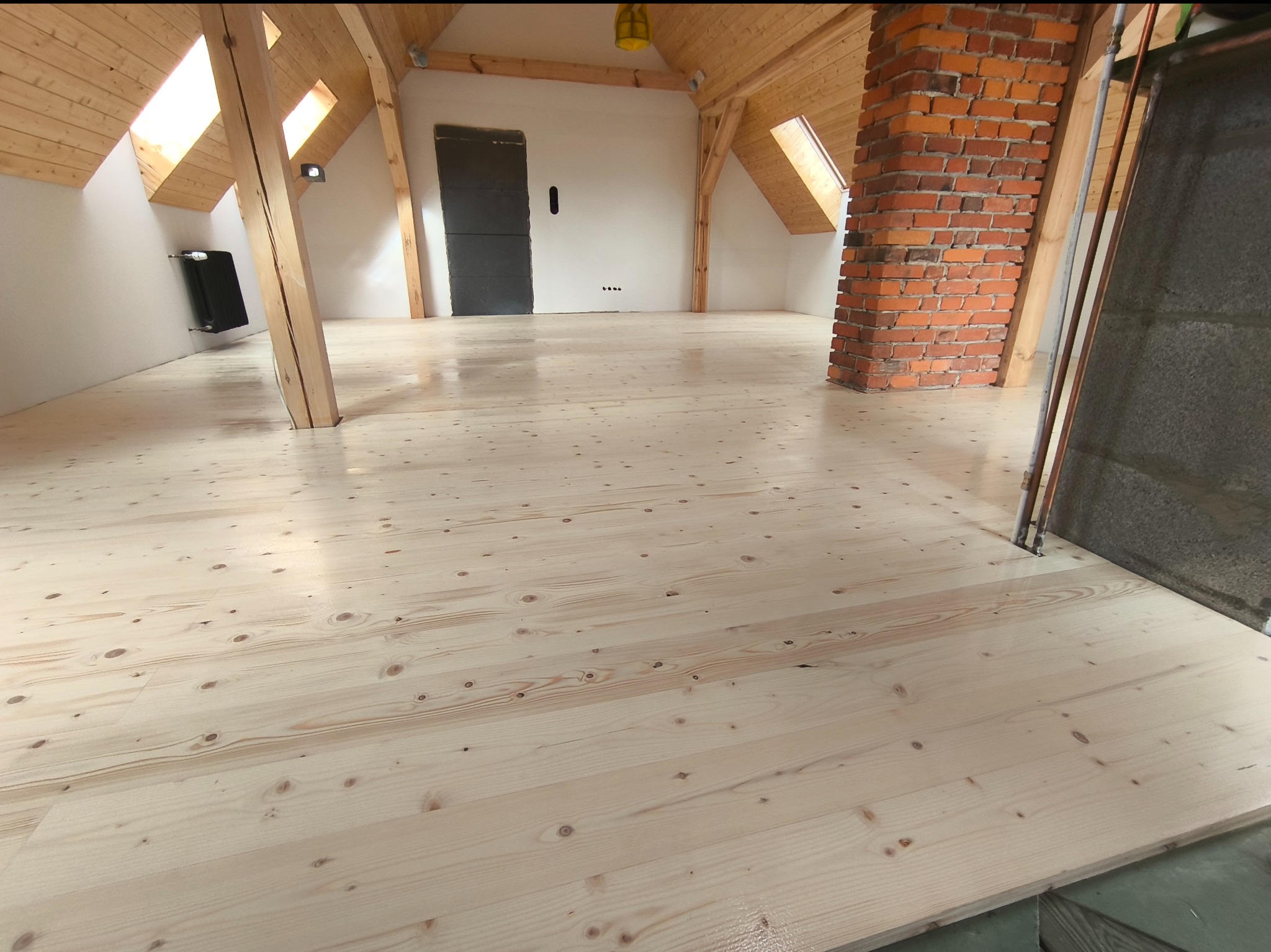 Jasna, odnowiona podłoga świerkowa w przestronnym, jasnym poddaszu, z widoczną strukturą drewna i charakterystycznymi sękami, po cyklinowaniu i lakierowaniu, z ceglanym kominem w tle.
