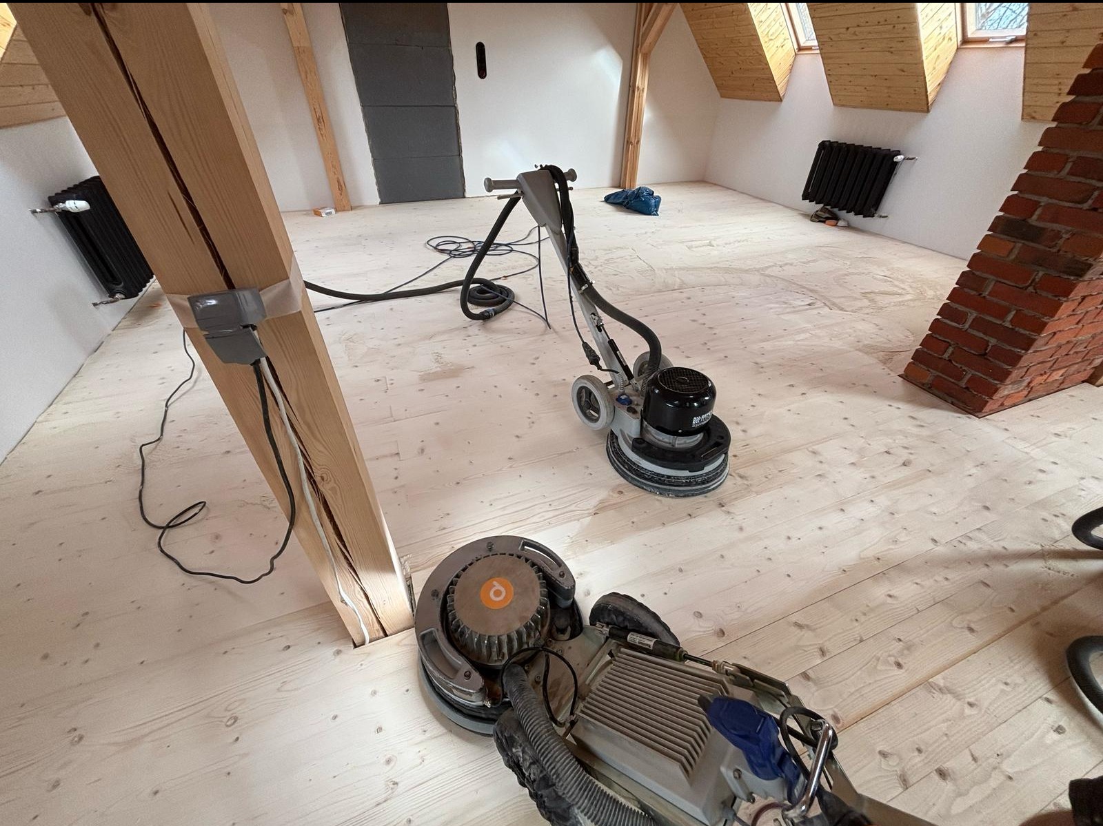 Dwa urządzenia do obróbki drewnianej podłogi w trakcie pracy w jasnym, przestronnym pomieszczeniu na poddaszu, widoczna drewniana belka konstrukcyjna i fragment komina z czerwonej cegły.