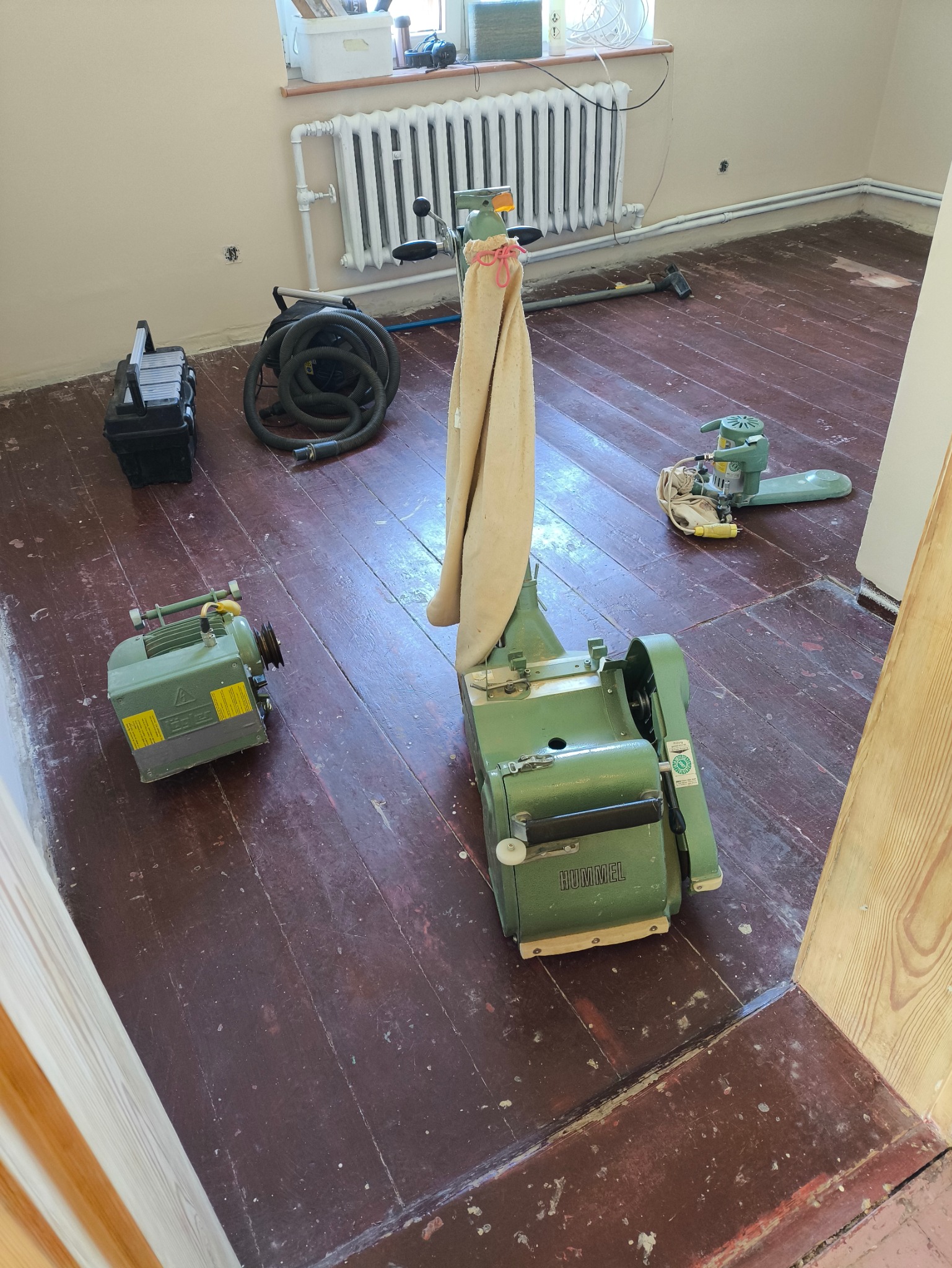 Parkiet przed odnowieniem: maszyna Hummel do cyklinowania, odkurzacz, szlifierka kątowa, silnik. Widoczne ślady starego lakieru i ubytki. Przygotowanie do prac.