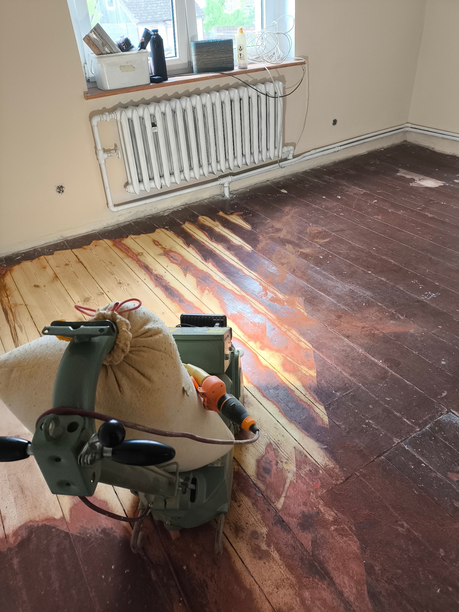 Cyklinowanie podłogi w trakcie realizacji. Część podłogi odnowiona, druga część w kolorze ciemnego brązu. Widoczna maszyna do cyklinowania.