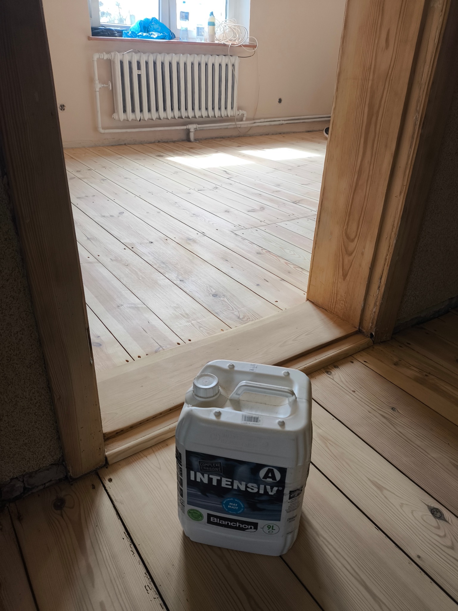 Jasna, odnowiona podłoga drewniana po cyklinowaniu, widoczny kanister z lakierem do parkietu Blanchon Intensiv. W tle grzejnik i okno. Widok zza futryny.
