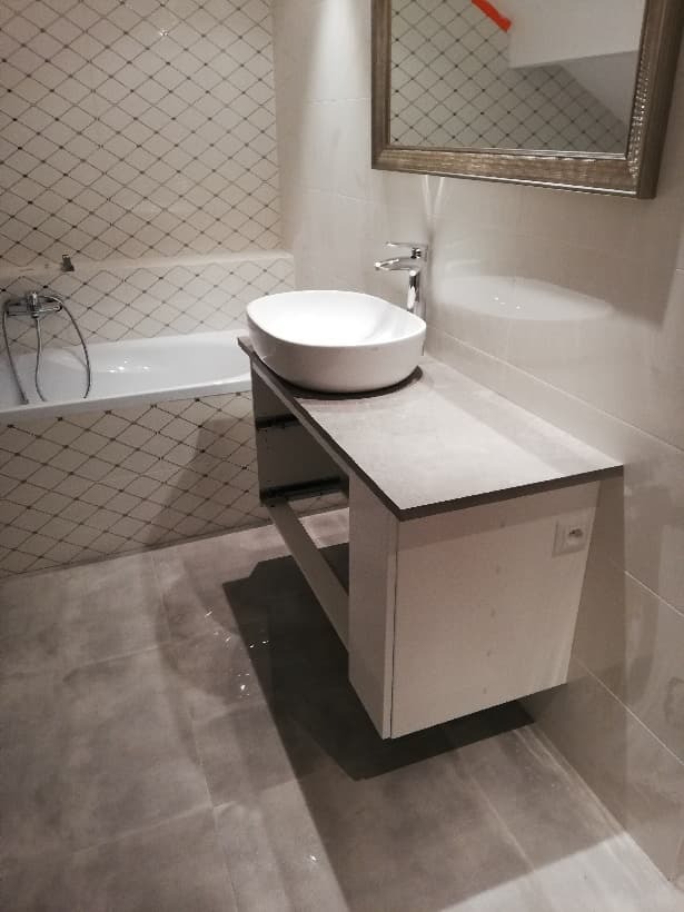 Nowoczesna łazienka z owalną umywalką nablatową, szarą płytą blatu i lustrem w ozdobnej ramie, widoczna część wanny z baterią i biało-szara mozaika na ścianie.