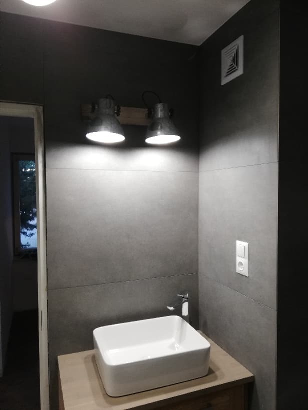 Nowoczesna łazienka z szarymi płytkami, białą prostokątną umywalką nablatową, chromowaną baterią i industrialnymi lampami na drewnianej listwie.