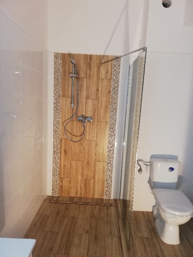 Kabina prysznicowa typu walk-in z drewnopodobną okładziną, mozaiką i odpływem liniowym w nowocześnie wykończonej łazience z białą ceramiką.