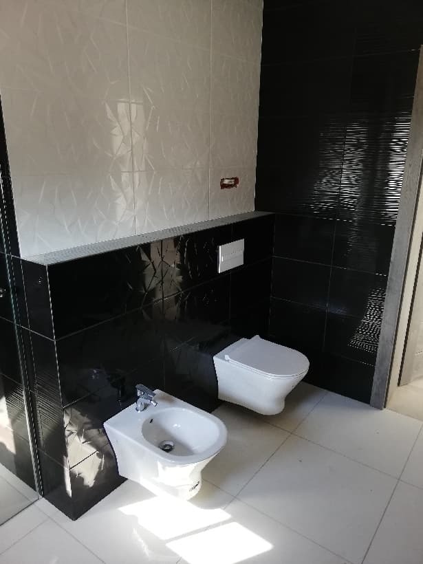 Nowoczesna łazienka z białymi i czarnymi płytkami, wiszącą toaletą i bidetem, biała podłoga z połyskiem.