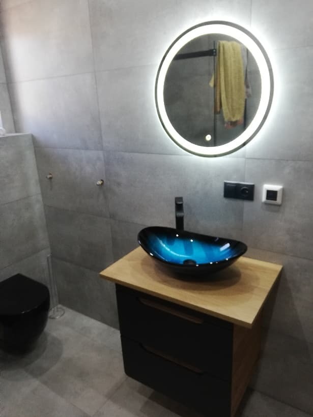 Nowoczesna łazienka z umywalką w kolorze niebieskim, okrągłym lustrem z podświetleniem LED i czarną toaletą w tle.