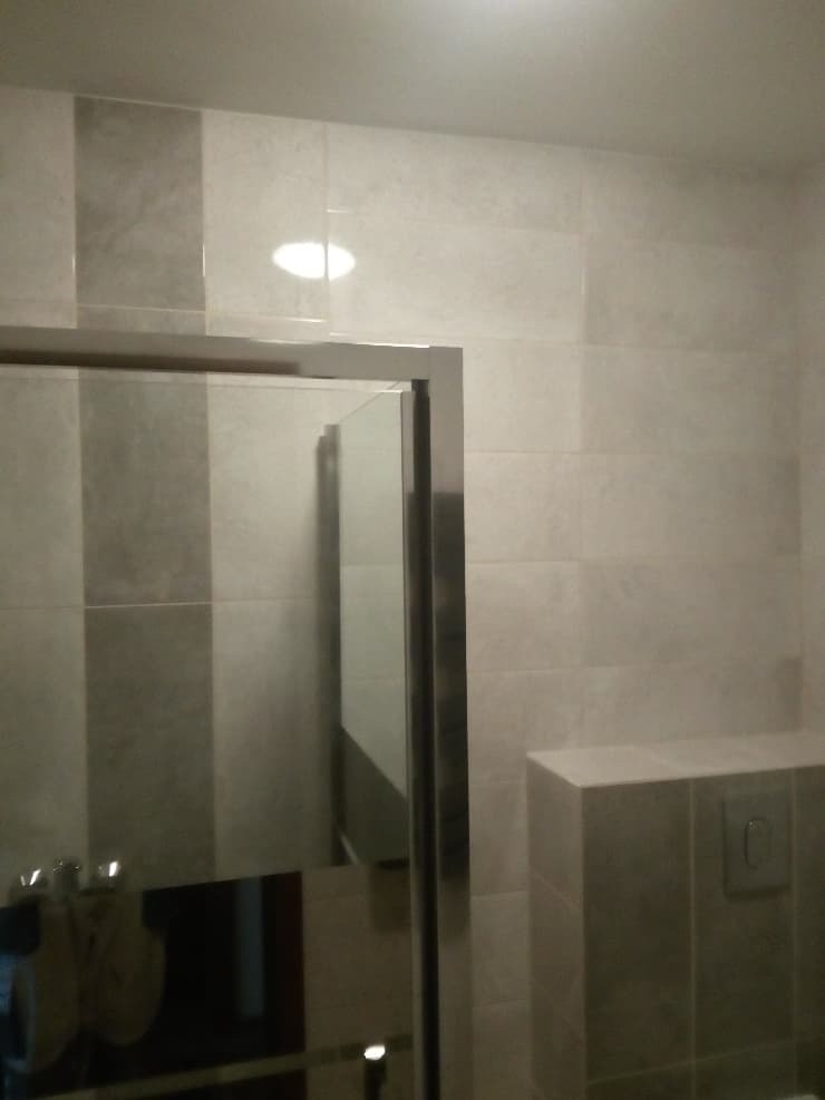 Fragment wykończonej łazienki z widoczną kabiną prysznicową z chromowanymi profilami i szklanymi drzwiami oraz częściowo widoczną zabudową WC z przyciskiem spłukującym, wykończoną płytkami...