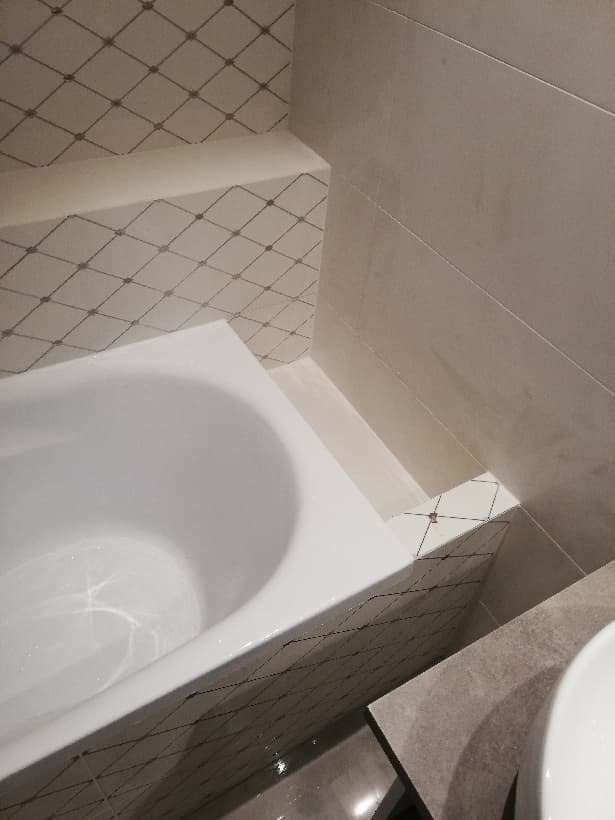 Nowa, biała wanna zabudowana płytkami z delikatnym wzorem siatki, połączona z jasnymi, podłużnymi płytkami ściennymi i elementem umywalki, w świeżo wykończonej łazience.