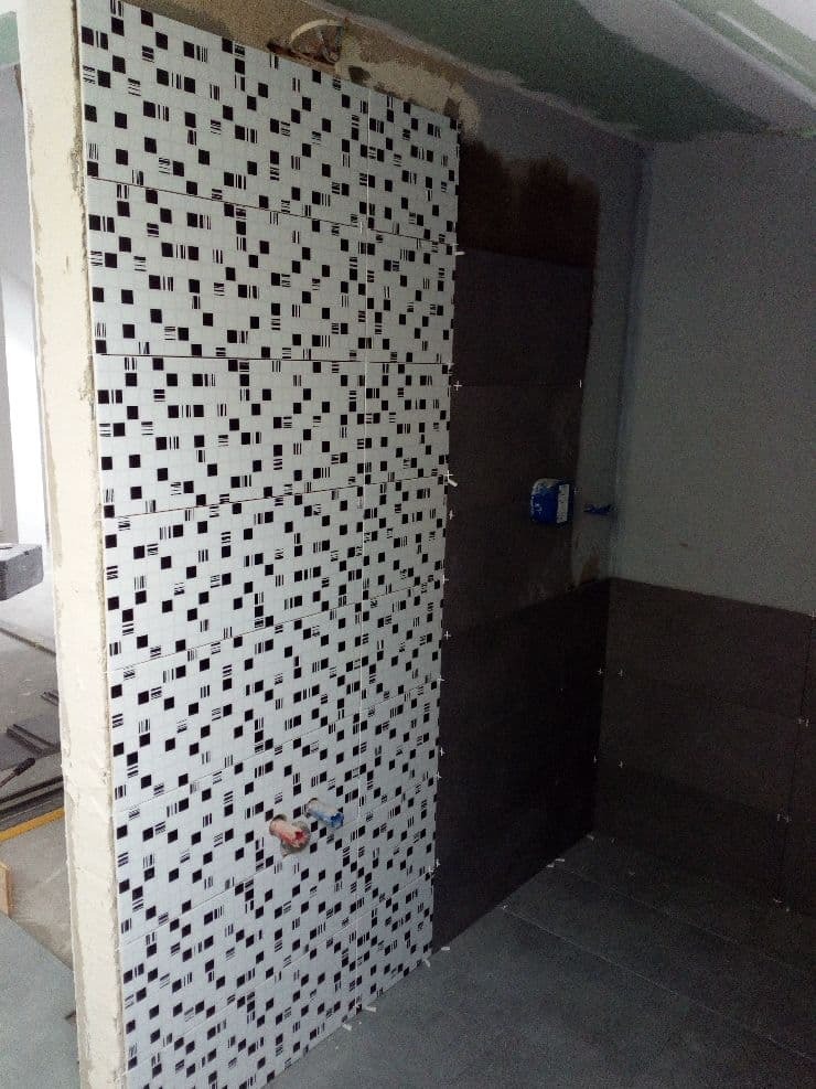 W trakcie układania białych płytek z czarnymi wzorami na ścianie łazienki, widoczne krzyżyki dystansowe i wystające rury instalacji wodnej.