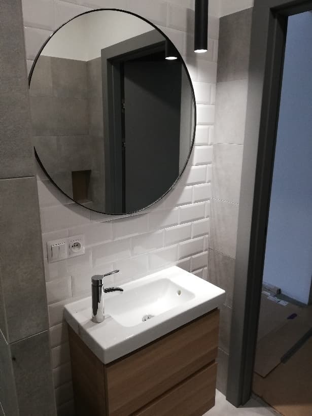 Nowoczesna łazienka z okrągłym lustrem w czarnej ramie, białą umywalką na drewnianej szafce, białą cegiełkową glazurą i szarą płytką na ścianie, widoczny fragment drzwi z szarą ościeżnicą.