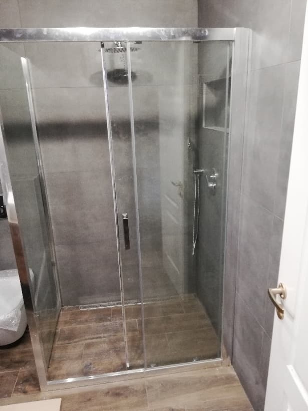 Kabina prysznicowa z przesuwnymi drzwiami, wykończona szarymi płytkami i drewnopodobną podłogą, widok z zewnątrz z częściowo widoczną toaletą w folii ochronnej.