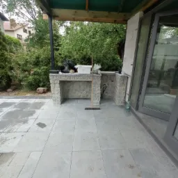 Zadaszony taras z kamienną zabudową grilla i szarymi płytami podłogowymi, widok na ogród i dom w tle.