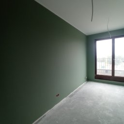 Ihor Kuzela - Pomalowane na oliwkowo ściany w pustym pokoju, surowa podłoga, wystające kable z sufitu, okno z brązową ramą.