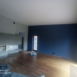 Ihor Kuzela - Wnętrze pokoju po remoncie z pomalowaną na granatowo ścianą, drewnianą podłogą, nowoczesnym kominkiem i oknem balkonowym. Na podłodze widoczne narzędzia malarskie.