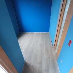 Ihor Kuzela - Świeżo pomalowane na niebiesko ściany i jasna drewniana podłoga w pustym pokoju, widoczne fragmenty ościeżnic drzwiowych i wystający kabel ze ściany.